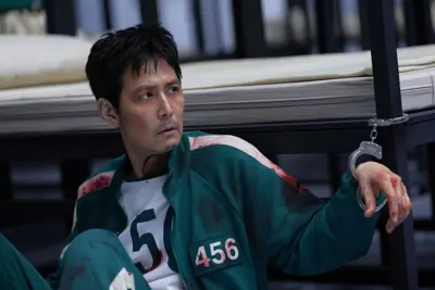 En man i grön träningsjacka med nummer 456, handfängslad vid en säng. Han har blod i ansiktet och på kläderna.