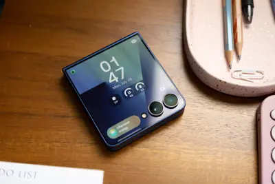 En hopfälld, blå mobiltelefon med två kameror ligger på ett träbord. Skärmen visar klockan 01:47.