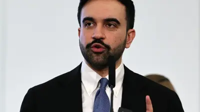 En man i kostym talar i en mikrofon. Han har mörkt hår och skägg.