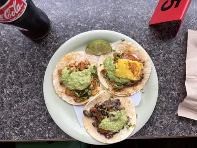 En tallrik med tre tacos med guacamole, limeklyfta och en Coca-Cola flaska.