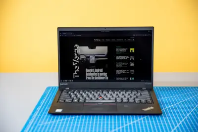 En svart Lenovo X1 Carbon laptop står öppen på ett blått skärmatta. Skärmen visar webbsidan The Verge.