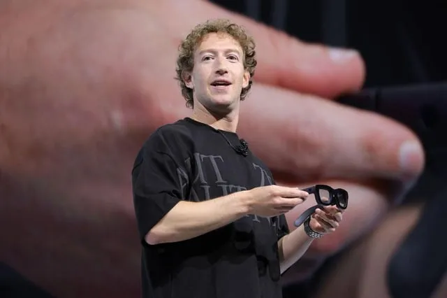 Mark Zuckerberg håller i ett par svarta glasögon på en scen. I bakgrunden syns en stor hand.