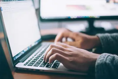 En person skriver på en laptop. En datorskärm syns i bakgrunden.